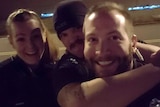 A police officer grabs another man around the neck and a female police officer stands behind. All three are smiling.