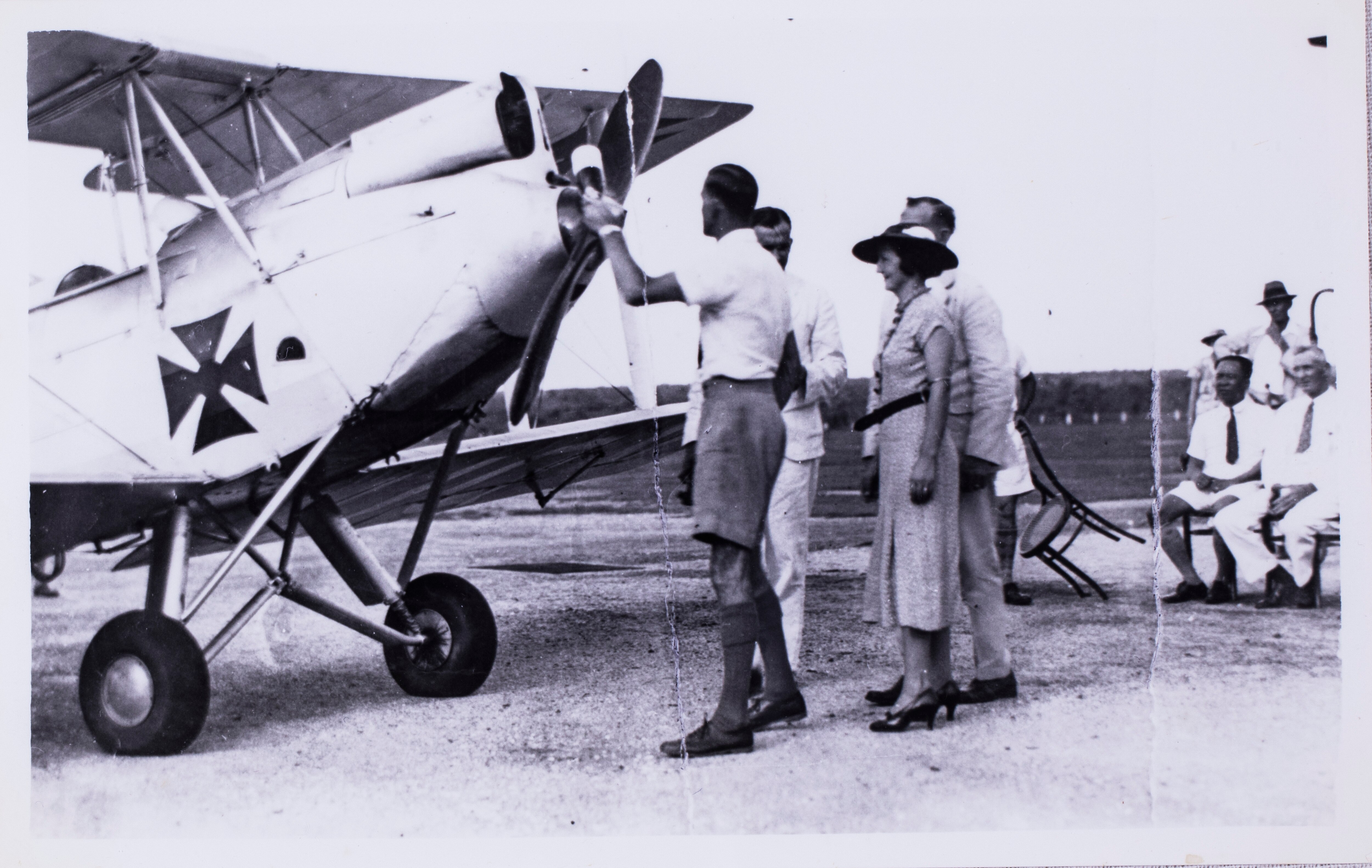 A man holds the propeller of an old plane while a group of people watch on.  The photo is old and in black and white.
