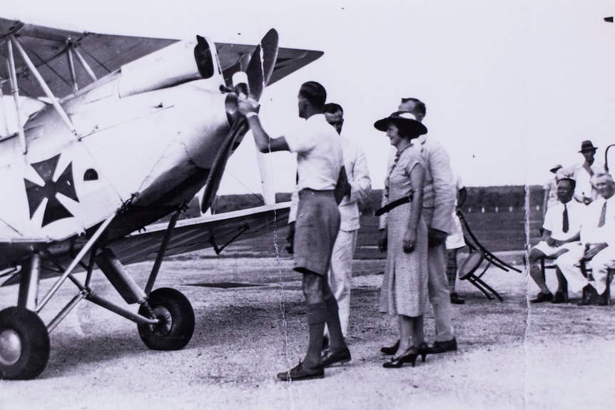 A man holds the propeller of an old plane while a group of people watch on.  The photo is old and in black and white.