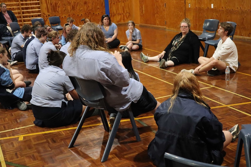 A group of high school students listen to Magda Szubanski who sits on the floor of a gym