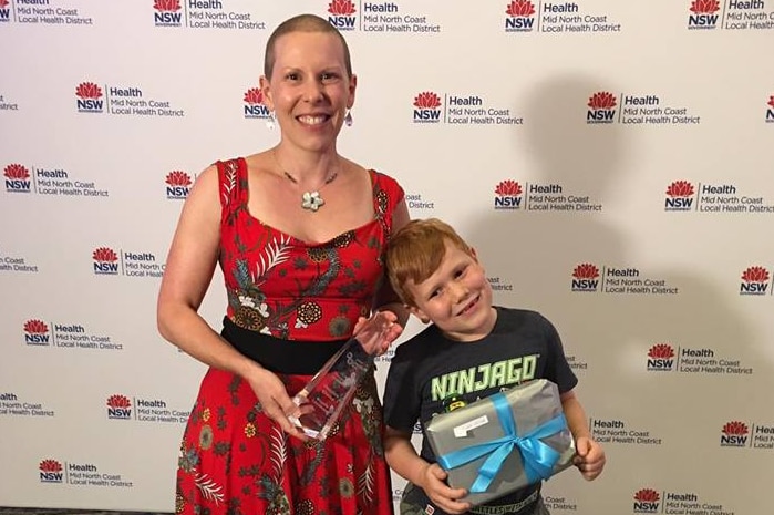 A woman stands with her arm around her son, holding an award.
