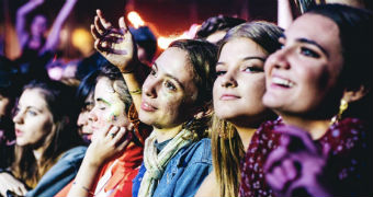 Five women stand in a row and smile and look to a stage as they listen to music at a festival.