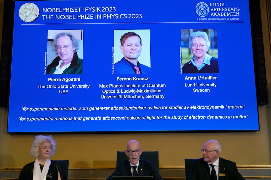 Two men and a woman are seated at a desk below a blue slide showing the faces and biographies of two other men and a woman.