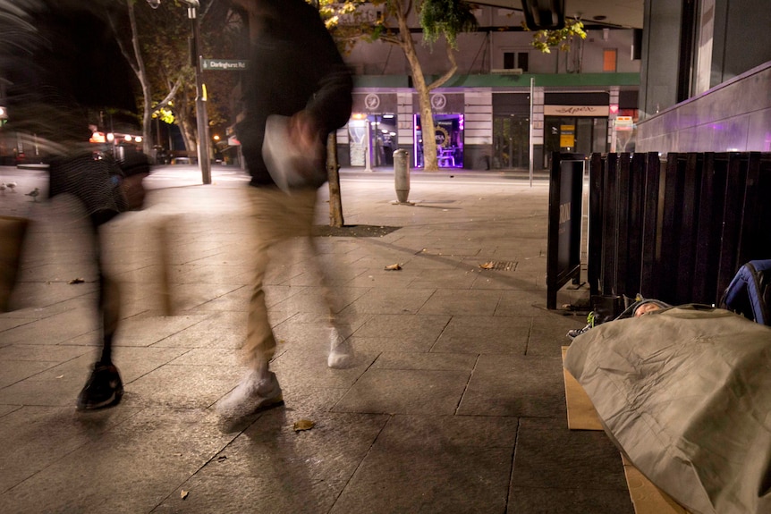 Two people walk past a homeless man sleeping in a swag on the ground