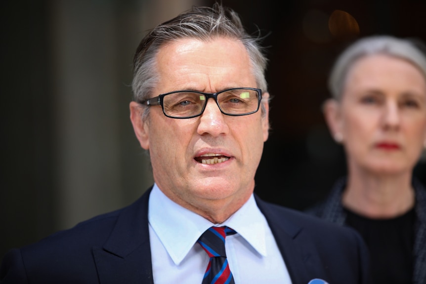 A man in a suit, tie and glasses speaks at a press conference.