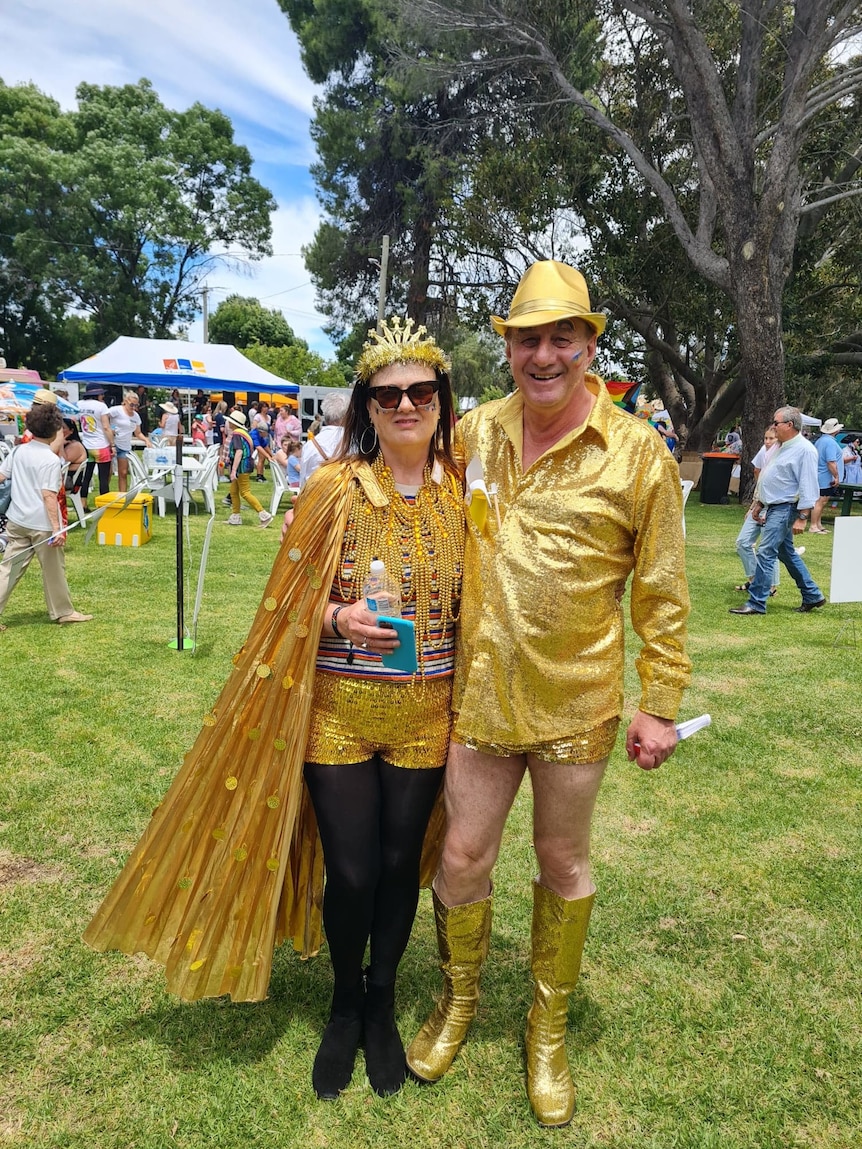 A woman in a sequin gold shirt and cape stands next to an older man in a gold sequin shirt, shorts and high heeled boots.
