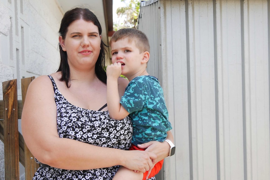 A woman in a summer dress standing in front of a gate, holding a young boy in her arms.