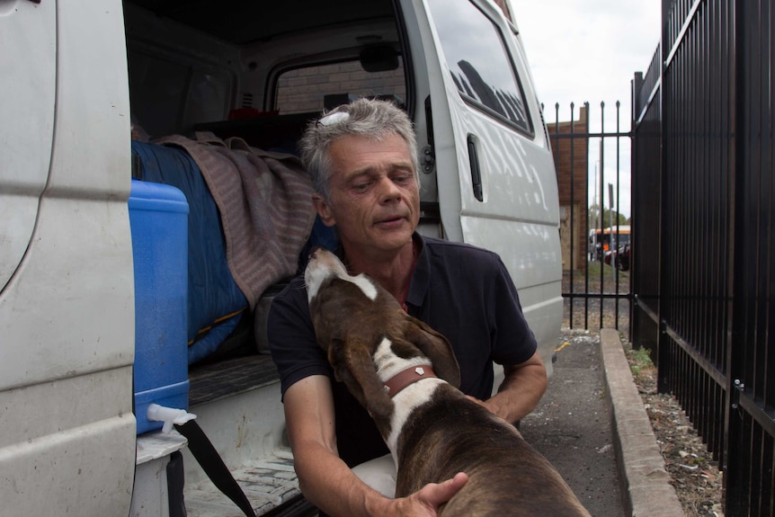 A man kneels next to a white van as a dog leans its head on his shoulder. Bed visible in van.