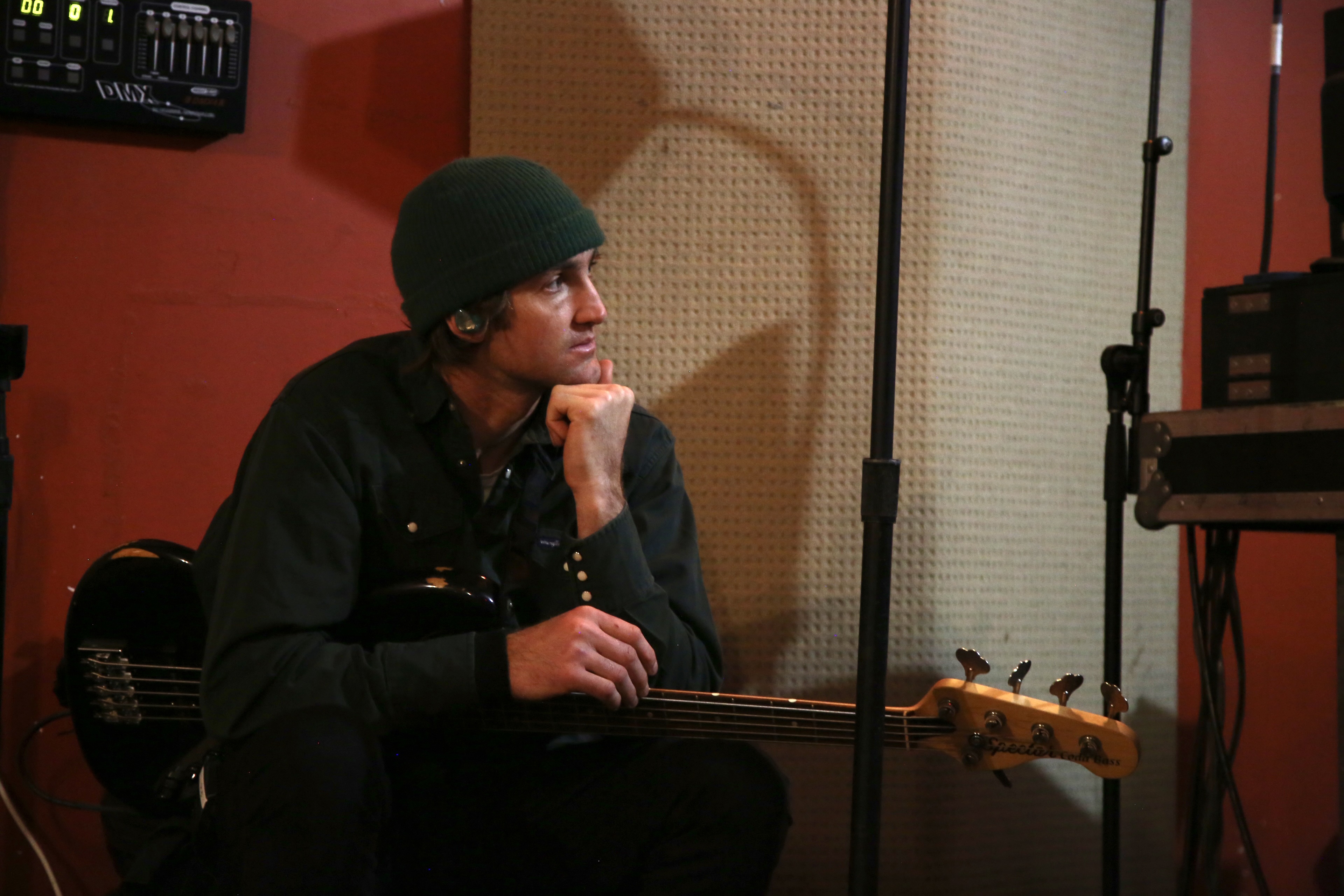 A man wearing a beanie sits on a couch holding a bass guitar, his hand resting under his chin
