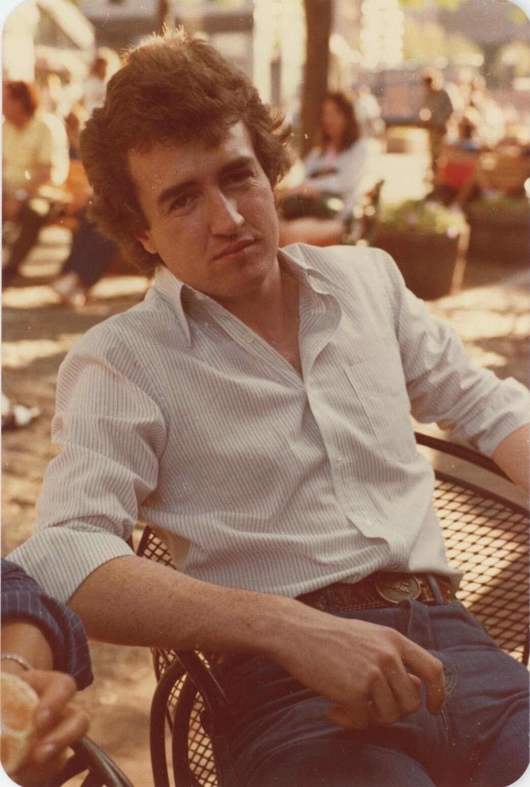 A young man sits in a chair in a courtyard in a white shirt and jeans in the 1970s