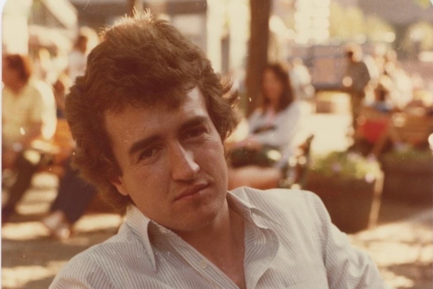 A young man sits in a chair in a courtyard in a white shirt and jeans in the 1970s