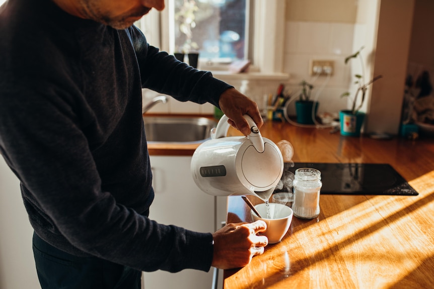 Man stands in dimly lit kitchen holding an electric kettle and pouring from it into a white mug.