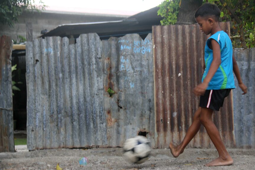 Young boy in an alley in front of a corrugated iron fence kicking a soccer ball.
