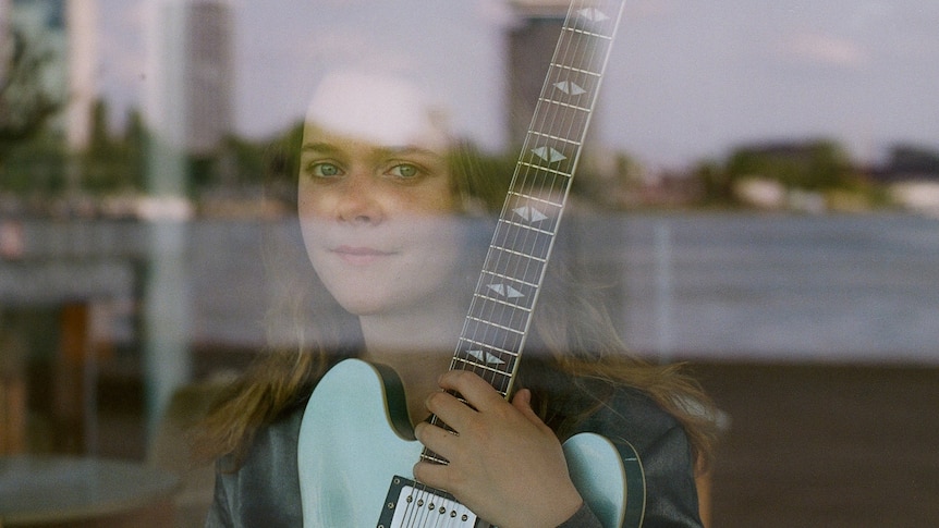 Ella Zirina holds a blue guitar, photographed through a reflective window