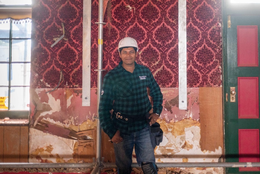 Man with white hard hat and tool belt leans on scaffold frame smiling