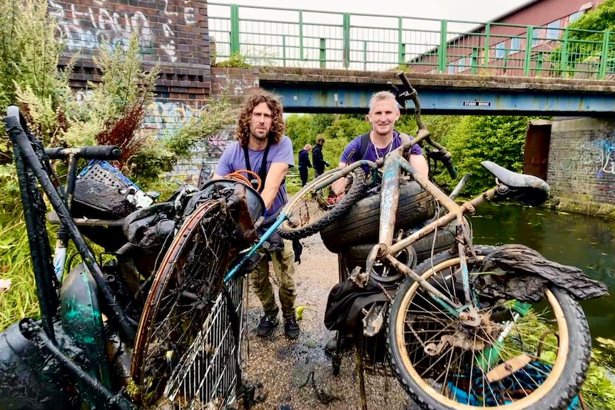 Two men, one with curly hair, another with grey hair, pose in a canal with rusted bikes.