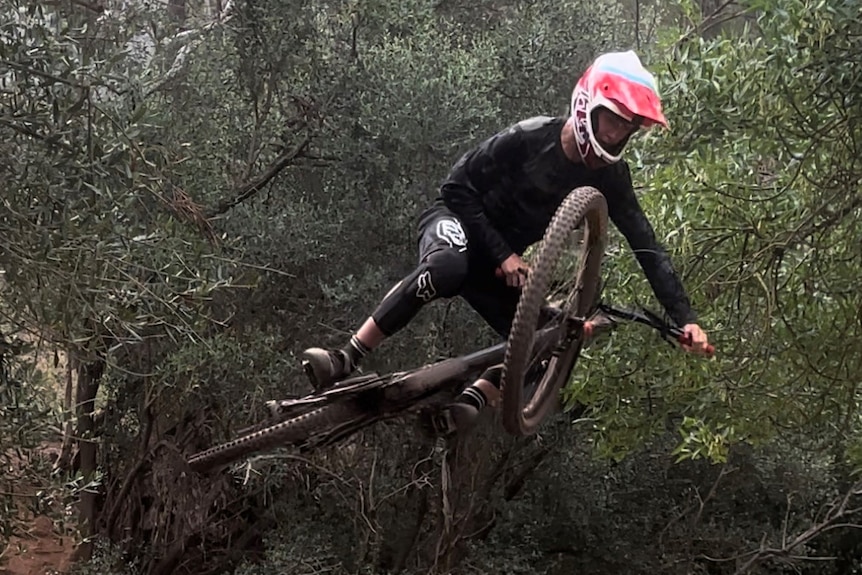 A teenager in protective bike riding gear is suspended in the air after going over a jump on his mountain bike. 