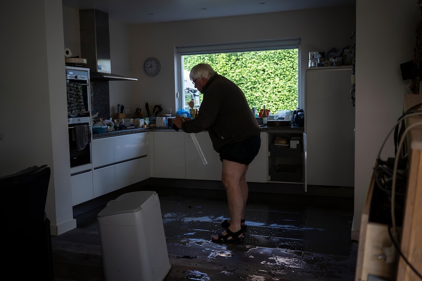 Un homme âgé se tient debout, sans porter de pantalon, dans sa cuisine dans des eaux de crue jusqu'aux chevilles. 