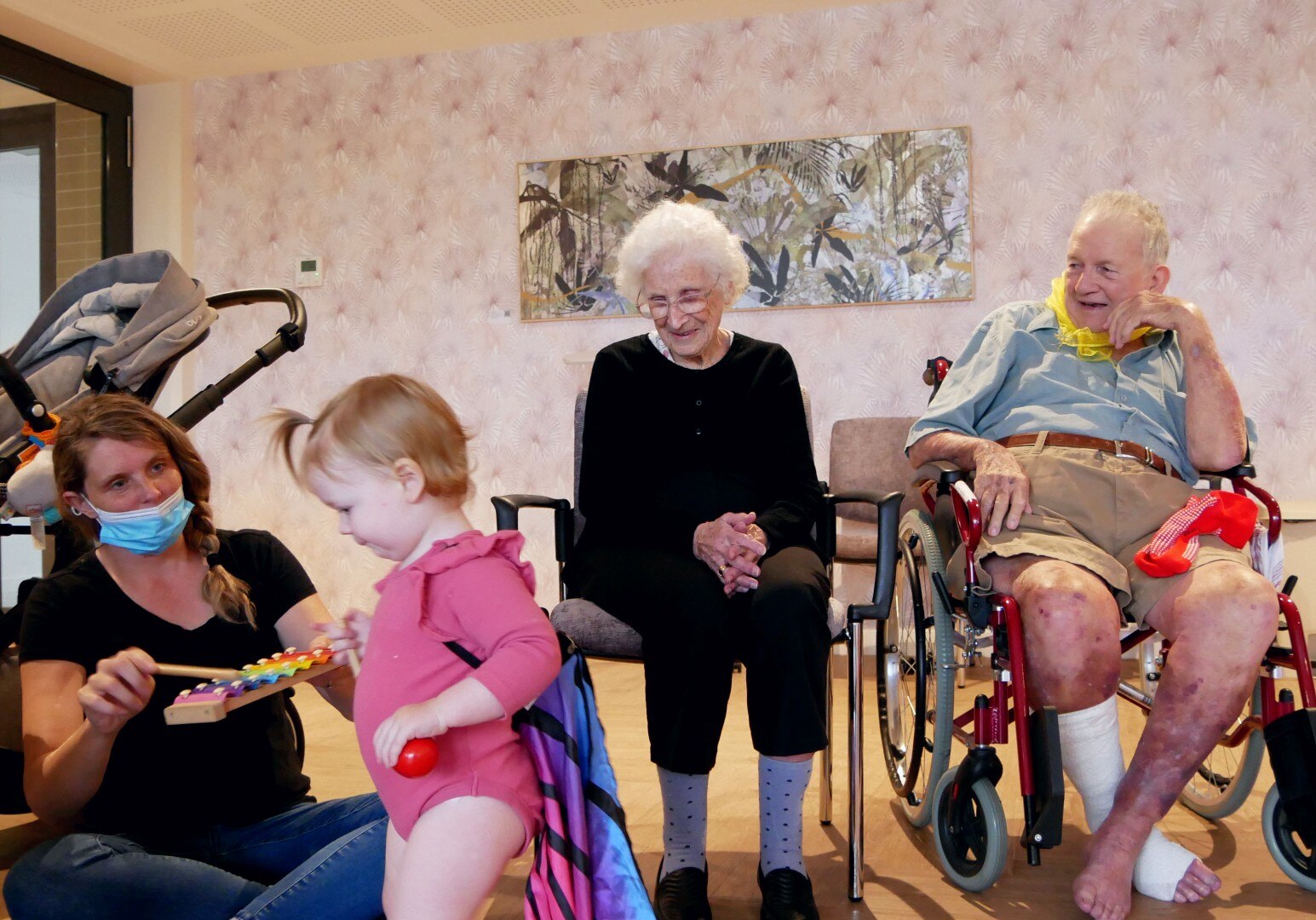 Elderly people sit in chairs, looking at a small toddler playing with a xylophone