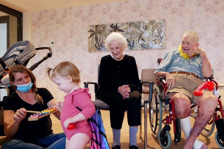 Elderly people sit in chairs, looking at a small toddler playing with a xylophone