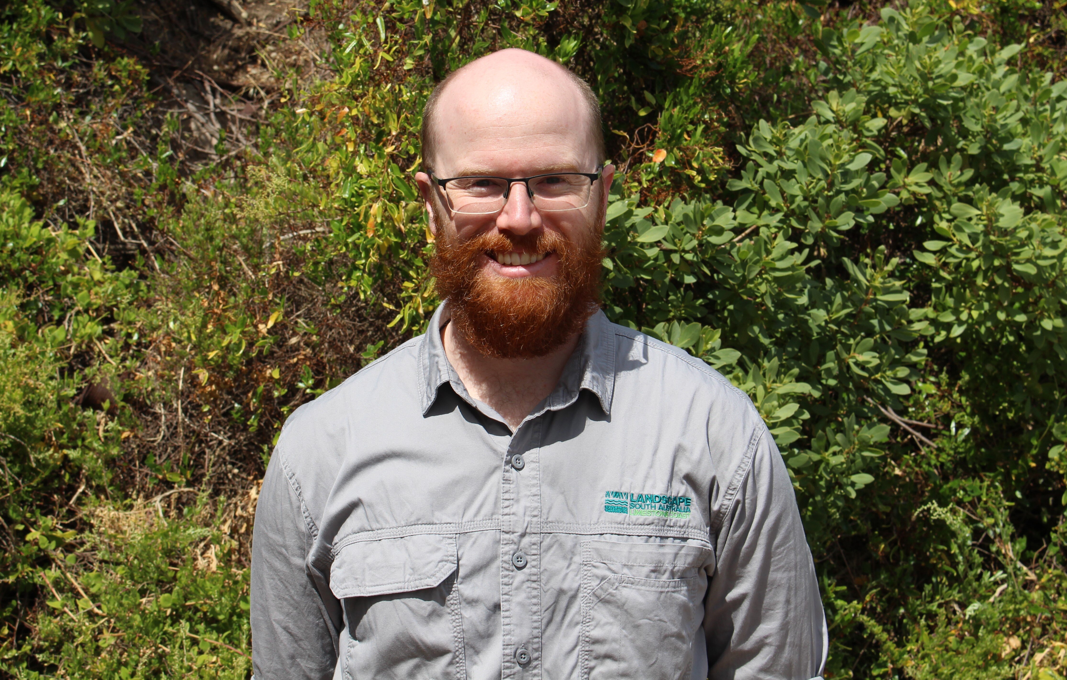 A bald man with a beard smiles at the camera with green bushes behind him