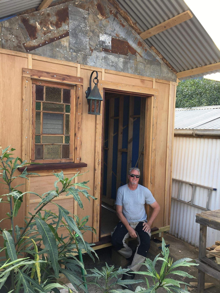 A man sits on the steps of a tiny house he is constructing in his backyard, smiling.
