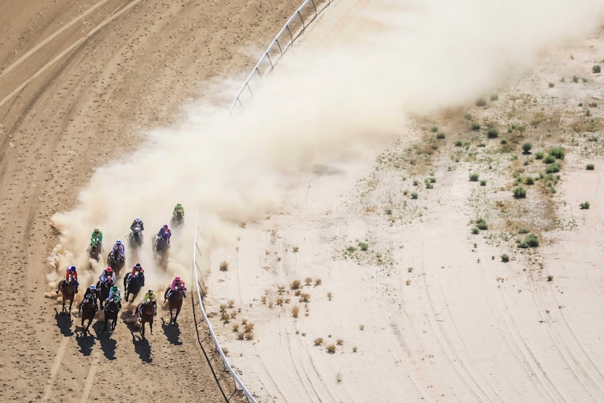 An aerial view of jockeys in colourful jerseys racing horses around a dirt racecours with a plume of dust rising from the track.
