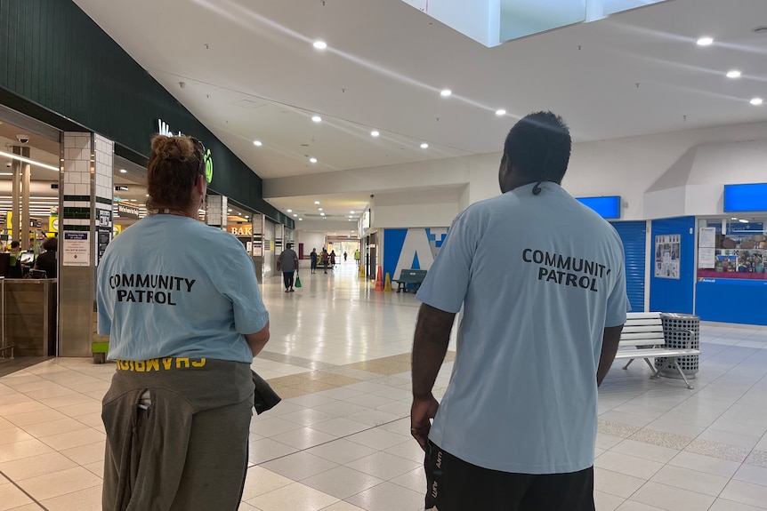 Two people stand in a shopping centre with their backs to the camera and shirts that have the words community patrol on them