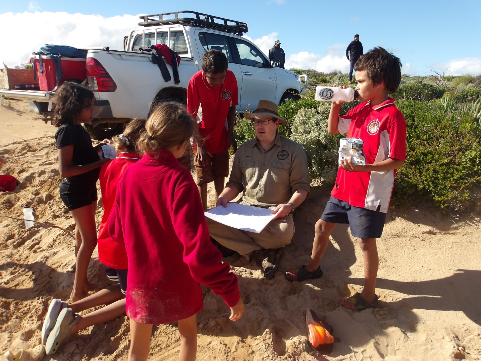 Students in red uniforms listen to Aaron Camens, who sits with a chart in front of him. A ute is in the background with eskies.
