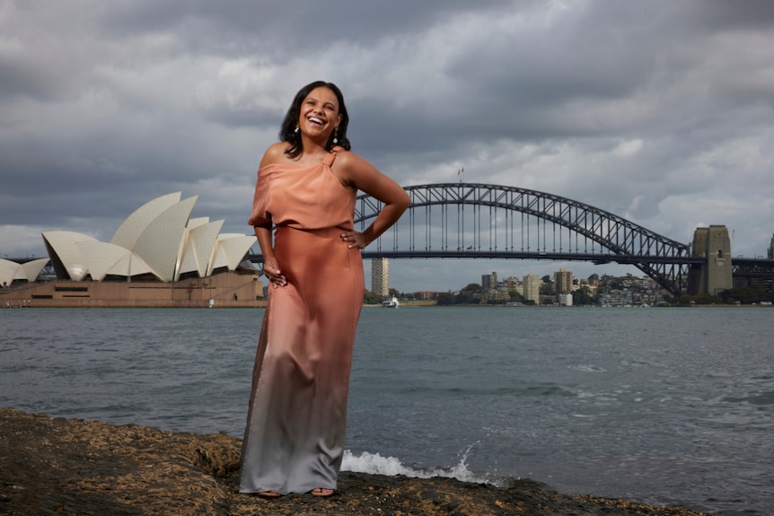 A smiling woman in a pink-blue gradient dress stands on a rock overlooking Sydney Harbour.
