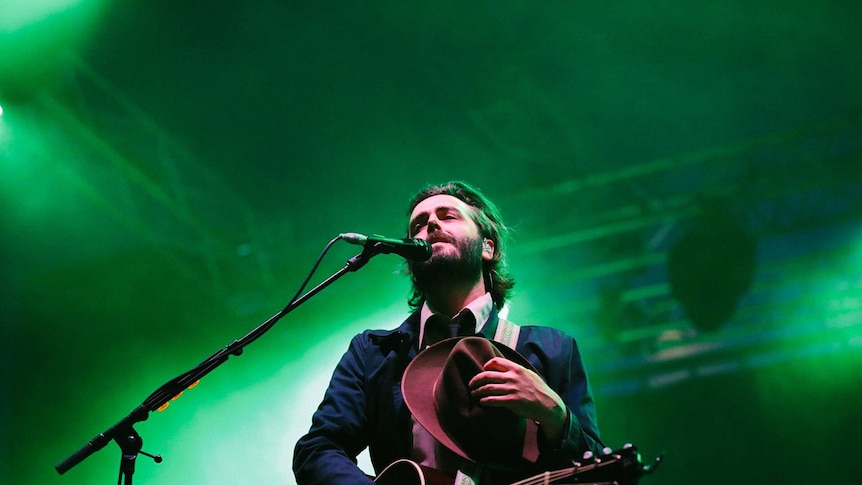Ben Schneider from Lord Huron wears a suit and tie while singing into a microphone. He's holding a hat and a guitar