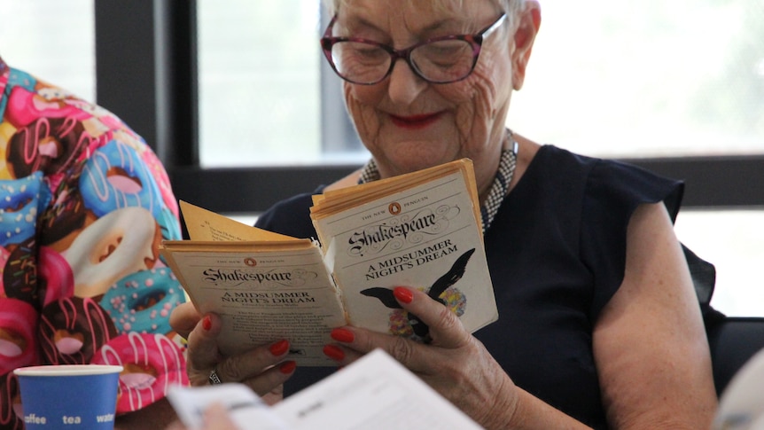 An older woman with glasses holds a copy of a midsummer night's dream which is cracking at the spine