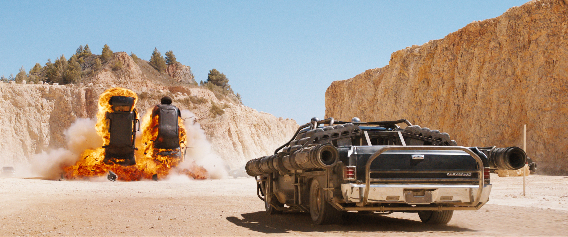 A souped up car sits in a dusty sandstone-lined landscape with two other cars burning mid-air in the distance.
