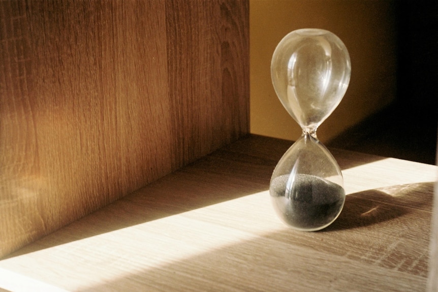 A photo of a glass hourglass sitting on a wooden desk in the sun
