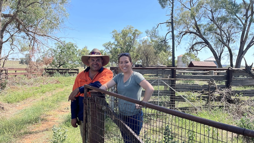 A man and woman stand, smiling, against a gate, with trees behind them.