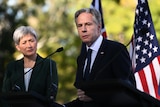 A white haired middle aged woman looks on as an older white haired man in suit speaking in front of flags on podium