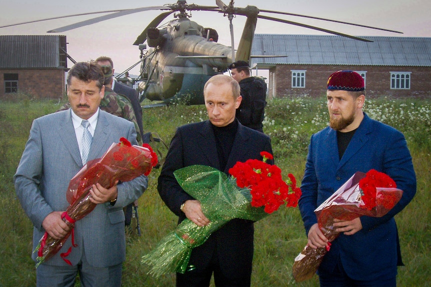 Vladimir Putin in a black turtleneck and suit clutches a huge bunch of red flowers, while standing with two men