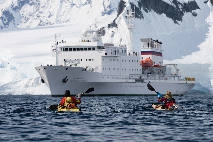 Sophie Ballagh and Ewan Blyth paddle their kayaks towards a ship. Behind it is a snow covered slope.