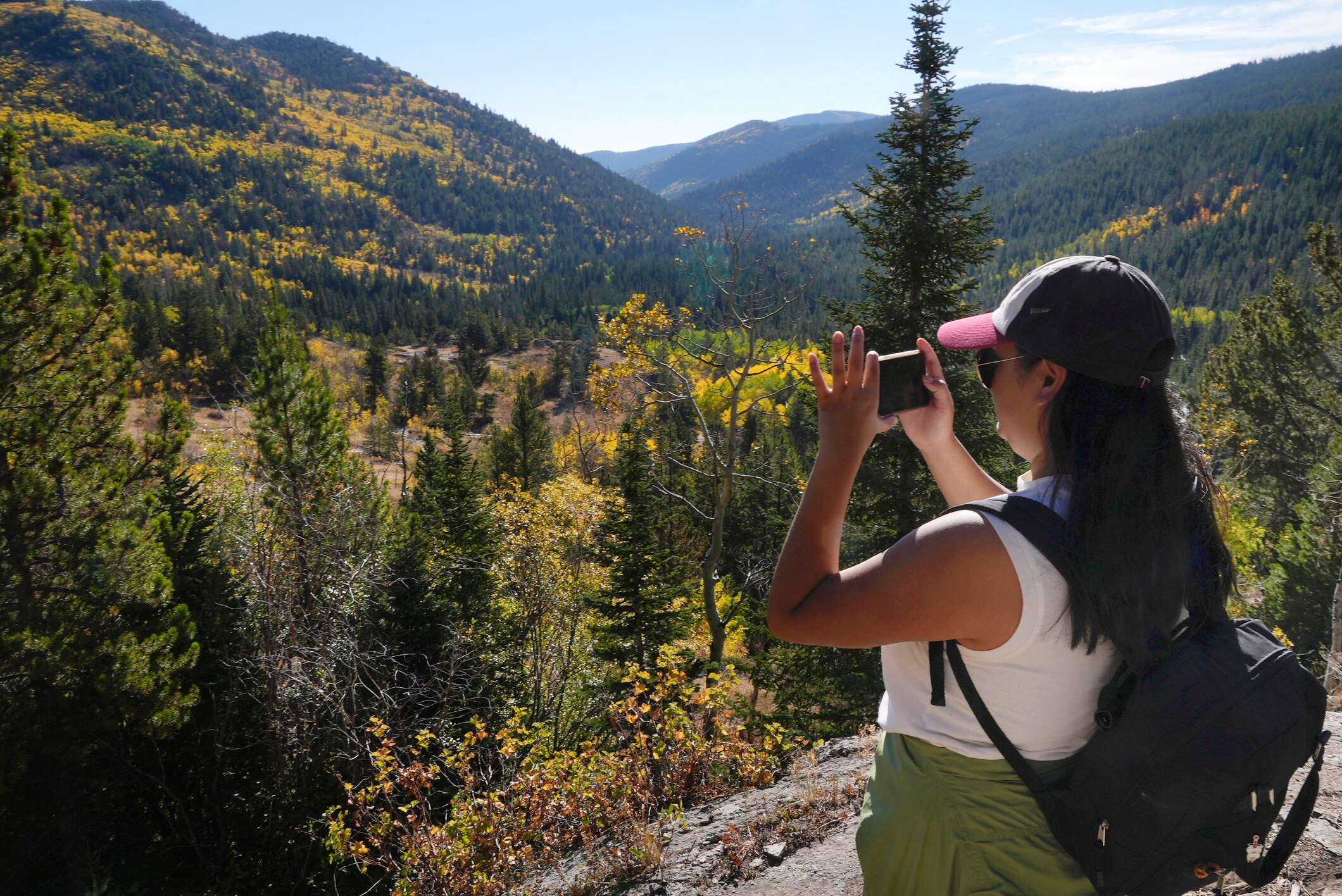 A woman wearing a cap, sunglasses and a backpack holds her phone up to photograph a view of a valley filled with trees.