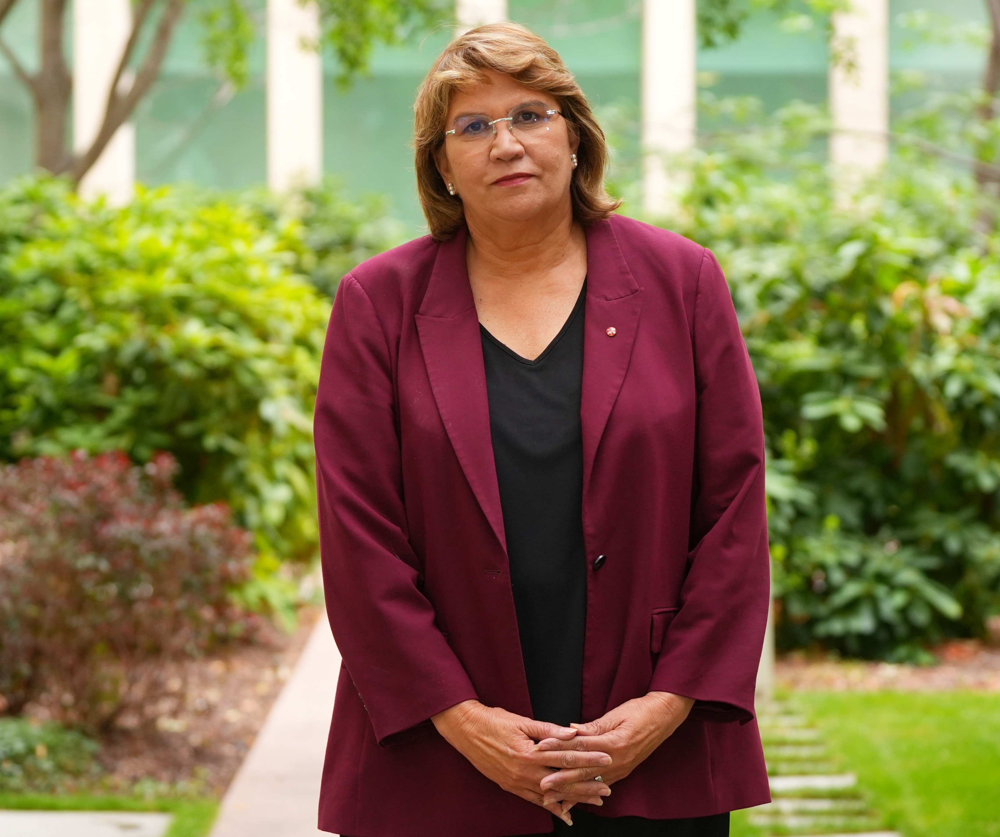 Kerrynne Liddle está de pie con las manos cruzadas frente a ella, vestida con una chaqueta granate y un top negro con una expresión neutra.