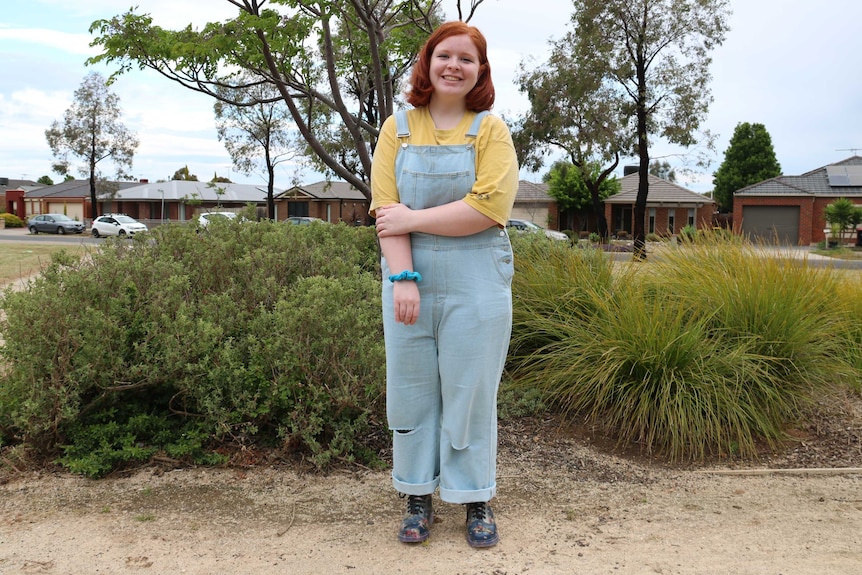 A young woman dressed in overalls stands outside in a suburban area