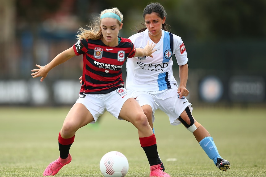 Two female soccer players, one wearing black and red stripes and another wearing white and blue, look at a ball during a game