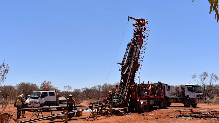 A drill rig in action on a mine set taking samples being operated by two men dressed in orange