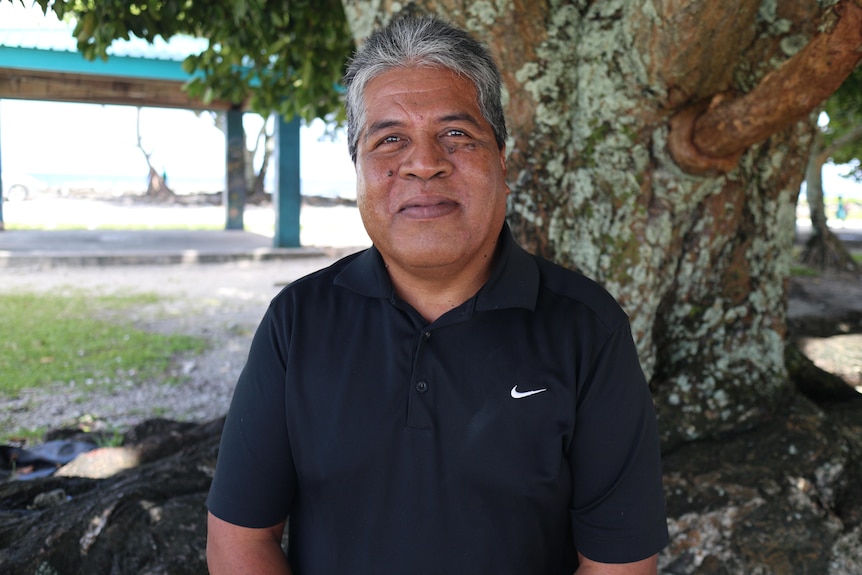 A smiling islander man wearing a dark polo shirt, salt and pepper hair, stands in front of a tree covered with moss.