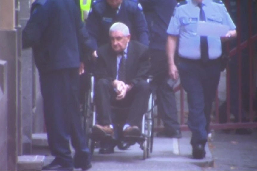 An older man in a wheelchair is escorted into court by police and security officers.