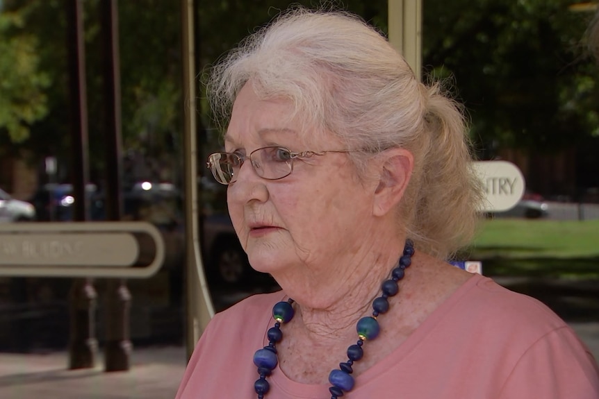 A woman with white hair in a ponytail, glasses, a pink tshirt and a blue necklace stands in front of glass doors