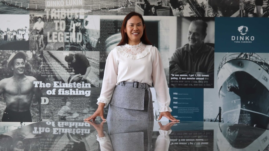 Woman standing with her reflection mirrored in a glass table, behind a wall collaged with pictures and newspaper articles.