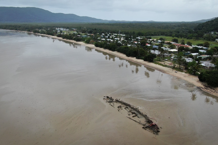 An aerial view with a grey coast to the left, a small series of houses and green mountains in the background. 