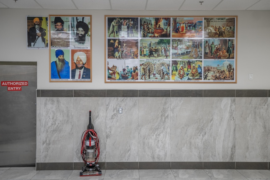 Photos of people, many wearing turbans, on a white and grey tiled wall. A vaccuum cleaner leans against the wall.