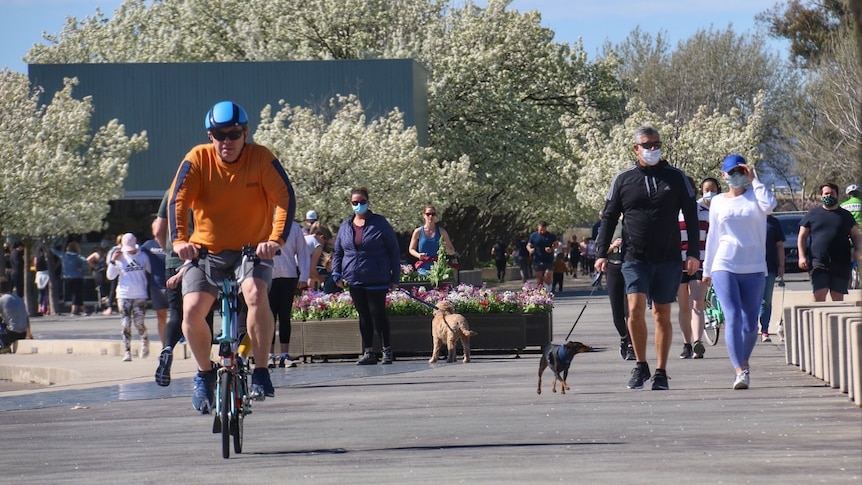 A man rides a bike in the foreground while masked people stroll behind him on a sunny boardwalk.